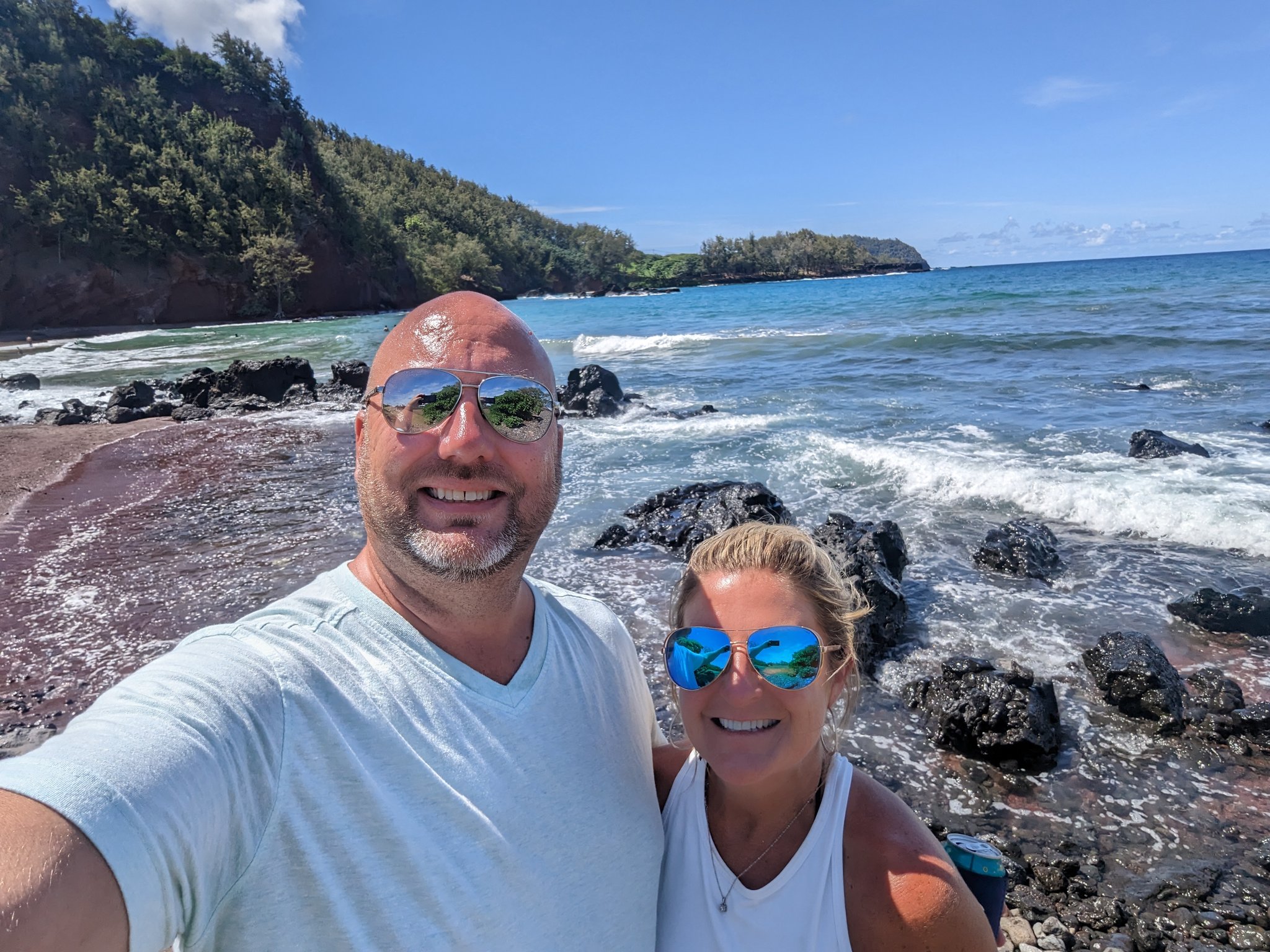 Kelli and me smiling for a selfie on a rocky shore with waves and green cliffs on the Road to Hana, Maui